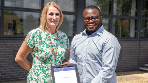 Two people standing in front of a building with large windows; one holds a framed certificate or award.